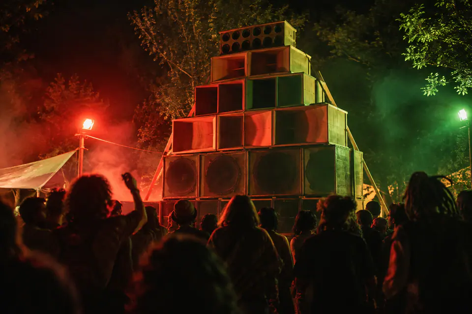 Un grand mur artisanal d’enceintes en bois domine des silhouettes qui dansent sous des lumières rouges et vertes dans un festival nocturne.